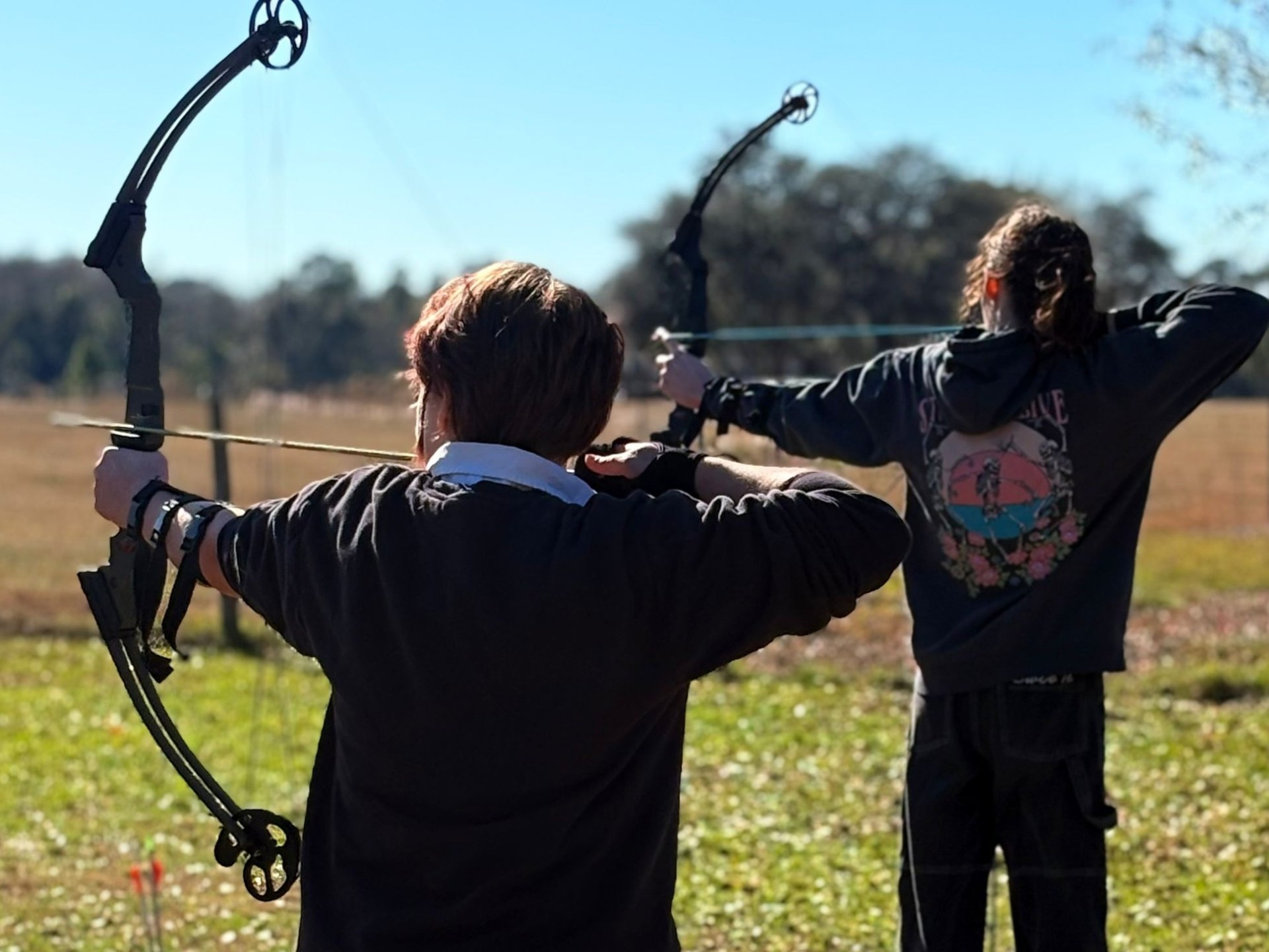 Two people practicing archery outdoors with compound bows.