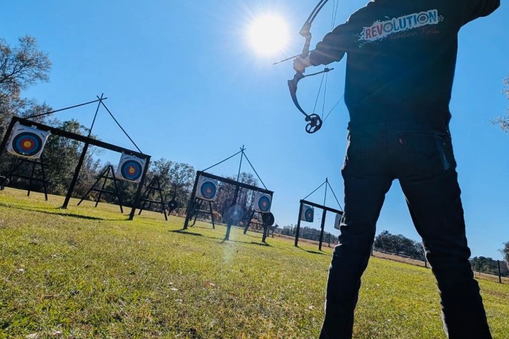 Person aiming a bow at archery targets in a sunny outdoor field.