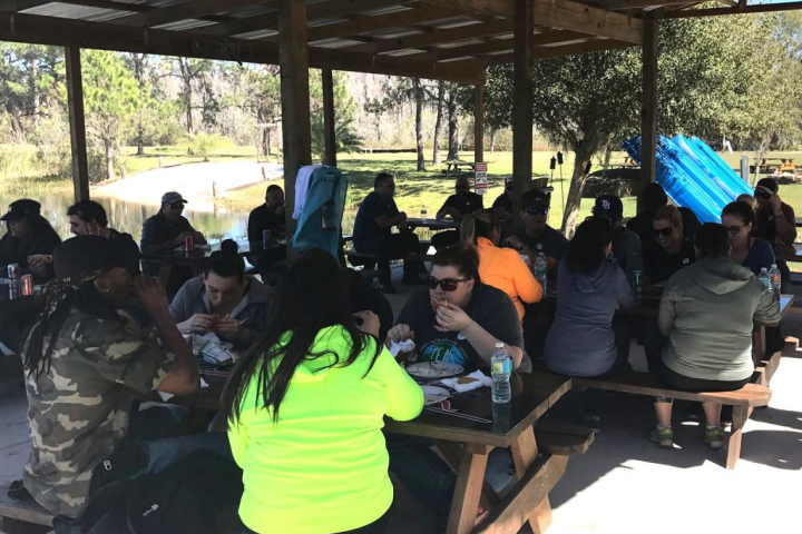 Group of people eating at outdoor picnic tables under a wooden roof.