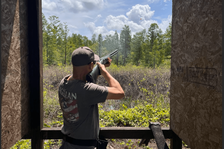 Person aiming a shotgun outside in a wooded area.