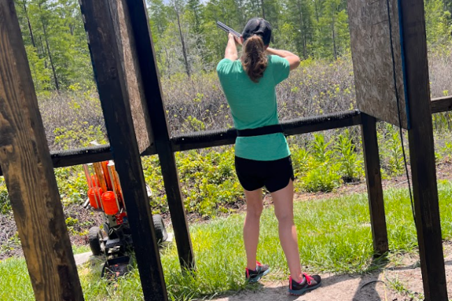 Person aiming a shotgun at an outdoor shooting range under a clear sky.