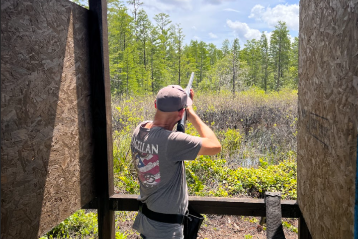 Person in a cap aiming a shotgun at flying clay target outdoors.