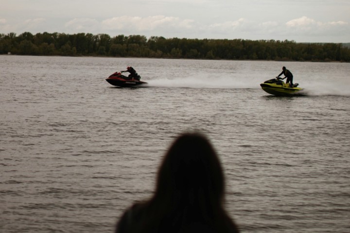 Person watches two jet skis on a lake under cloudy sky.