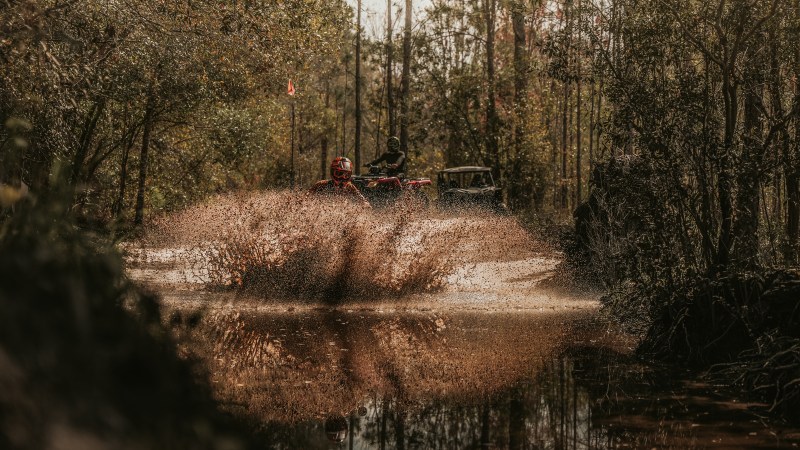 ATV splashing through muddy water in a forested area.