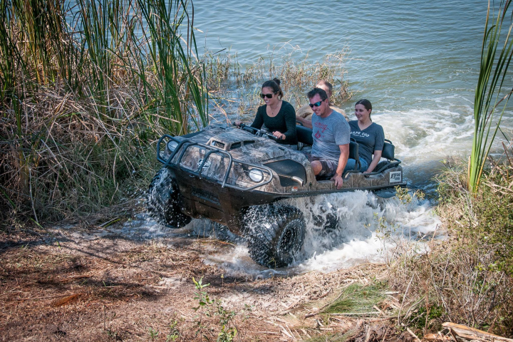 Four people riding an amphibious vehicle out of the water onto grassy land.