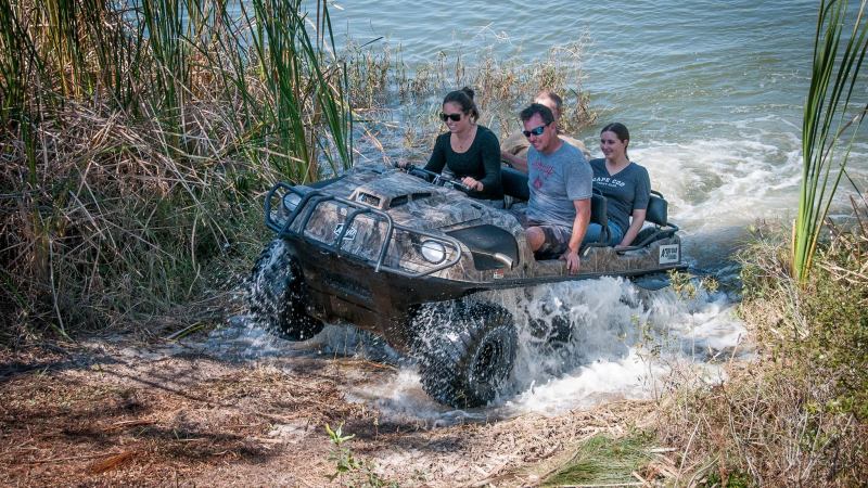 Four people riding an amphibious vehicle out of the water onto grassy land.