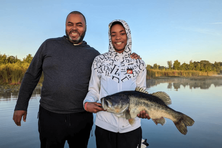 Two people smiling, holding a large fish by a lake.