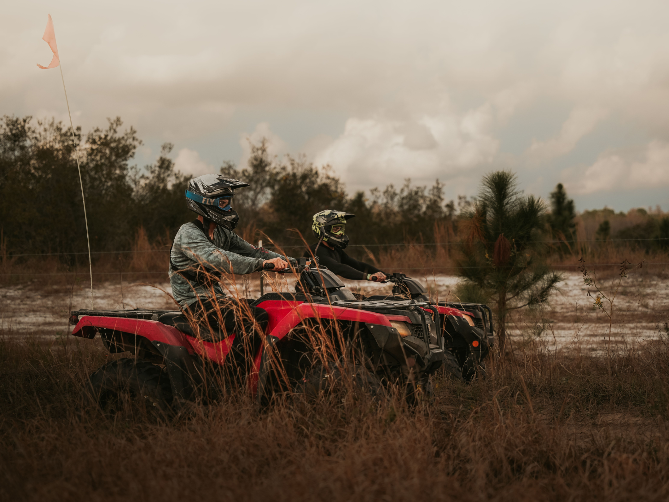Two people riding ATVs through tall grass on a cloudy day.