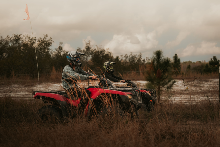 Two people riding ATVs through tall grass on a cloudy day.