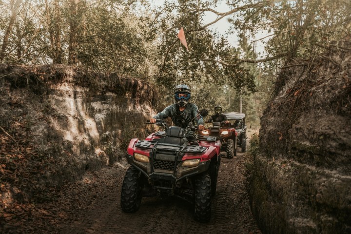 People riding ATVs on a dirt trail through a wooded area.
